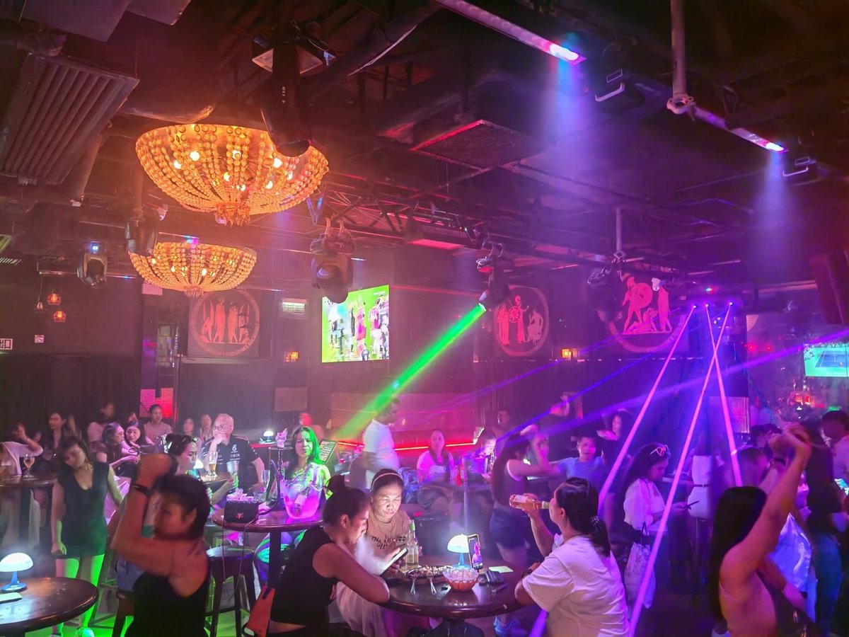 Vibrant Taboo nightclub interior with neon signs, golden chandeliers, purple and green lighting, laser beams, and crowds of people dancing and socializing on the dance floor